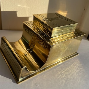 Puede incluir: Un organizador de escritorio vintage de latón, con un portalápices en forma de libro y una caja de dos niveles. El latón presenta intrincados diseños florales y geométricos grabados. El portalápices está abierto y las cajas tienen tapas.