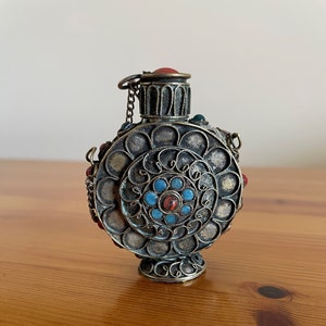 Puede incluir: Una pequeña botella redonda de color plateado con un diseño decorativo y piedras preciosas de colores. La botella tiene una cadena unida a la parte superior y un pequeño tapón redondo.