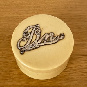 May include: A round, cream-colored trinket box with a silver-toned metal inscription that reads "Pin". The box has a lid and a base, and the inscription is in a decorative font. The box is sitting on a wooden surface.