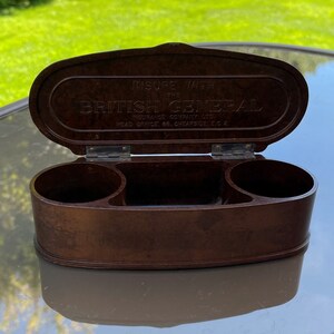 May include: A brown, oval-shaped, bakelite box with a hinged lid. The lid has the text "Insure with the British General Insurance Company Ltd. Head Office, 66, Cheapside, E.C.2."
