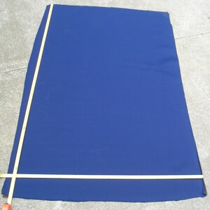 May include: A large, dark blue fabric rectangle is laid out on a concrete surface. A yellow measuring tape is laid across the fabric, with the end of the tape extending off the edge of the fabric.