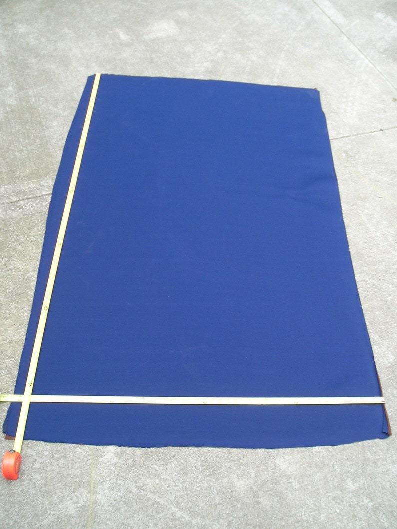 May include: A large, dark blue fabric rectangle is laid out on a concrete surface. A yellow measuring tape is laid across the fabric, with the end of the tape extending off the edge of the fabric.
