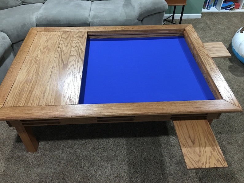 May include: A wooden coffee table with a blue felt game board insert and two pull-out side panels. The table has a natural wood finish and a rectangular shape.