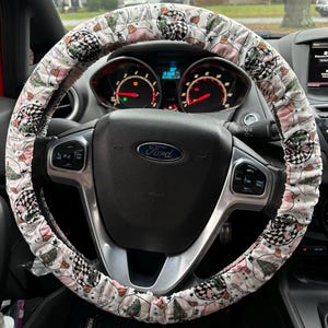 May include: A white steering wheel cover with a pink and green Christmas tree pattern. The cover has a checkered pattern and a white border. The cover is on a black steering wheel with a Ford logo.