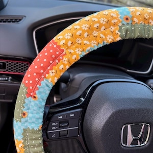 K&ouml;nnte beinhalten: Ein Lenkradbezug aus Stoff mit Patchwork-Design, bestehend aus abwechselnden Abschnitten mit senfgelbem Blumenmuster, hellblauem Vichy-Karo, orangefarbenem Stoff mit wei&szlig;en Punkten und olivgr&uuml;nem Stoff mit wei&szlig;en Sprenkeln auf einem schwarzen Lenkrad.