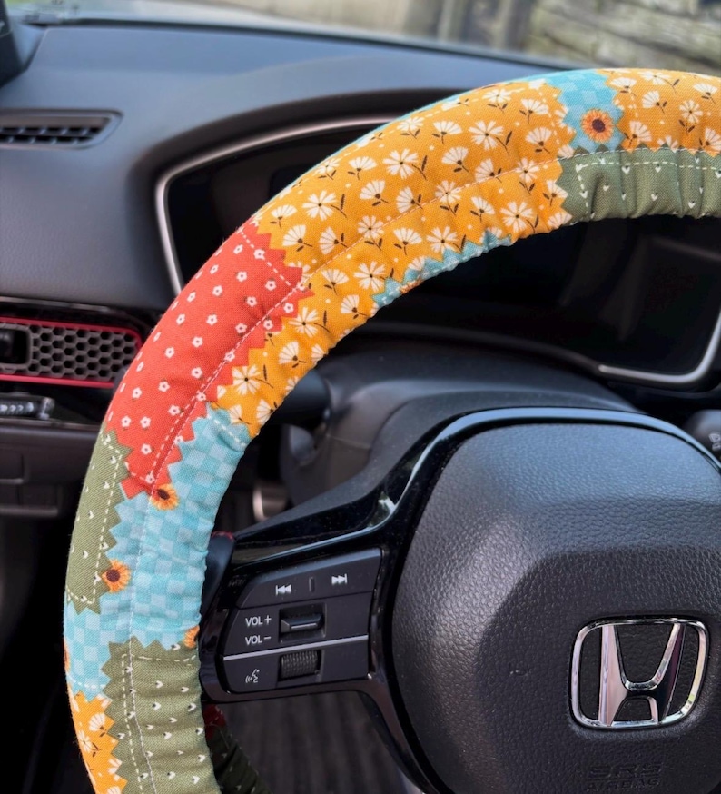 K&ouml;nnte beinhalten: Ein Lenkradbezug aus Stoff mit Patchwork-Design, bestehend aus abwechselnden Abschnitten mit senfgelbem Blumenmuster, hellblauem Vichy-Karo, orangefarbenem Stoff mit wei&szlig;en Punkten und olivgr&uuml;nem Stoff mit wei&szlig;en Sprenkeln auf einem schwarzen Lenkrad.