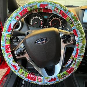 May include: A festive steering wheel cover featuring a Grinch-themed design. The cover is primarily light blue, red, and green, with images of the Grinch and text that reads "Grinchmas Days". The cover is wrapped around a black steering wheel with a Ford logo.