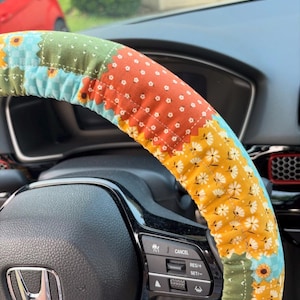 K&ouml;nnte beinhalten: Ein Lenkradbezug aus Stoff mit Patchwork-Design, bestehend aus verschiedenen Blumen- und geometrischen Mustern in Orange, Gelb, Gr&uuml;n und Hellblau, montiert auf einem schwarzen Lenkrad mit einem silbernen Honda-Logo in der Mitte.