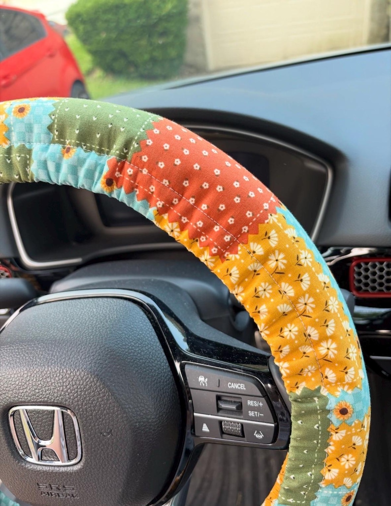K&ouml;nnte beinhalten: Ein Lenkradbezug aus Stoff mit Patchwork-Design, bestehend aus verschiedenen Blumen- und geometrischen Mustern in Orange, Gelb, Gr&uuml;n und Hellblau, montiert auf einem schwarzen Lenkrad mit einem silbernen Honda-Logo in der Mitte.