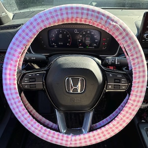 May include: A pink and white gingham steering wheel cover on a black steering wheel. The cover is made of fabric and has a classic checkered pattern. The car's dashboard is visible in the background.