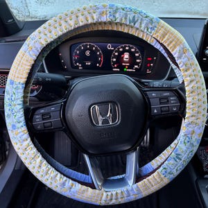 May include: A floral patterned steering wheel cover in shades of yellow, blue, and white. The cover is made of fabric and is wrapped around a black steering wheel. The car's dashboard is visible in the background.