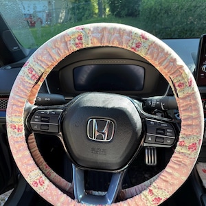 Op de afbeelding: Een roze bloemen stuurwielhoes op een zwart stuurwiel. De hoes heeft een herhalend patroon van roze rozen en groene bladeren op een lichtroze achtergrond. Het dashboard en de bedieningselementen van de auto zijn zichtbaar.