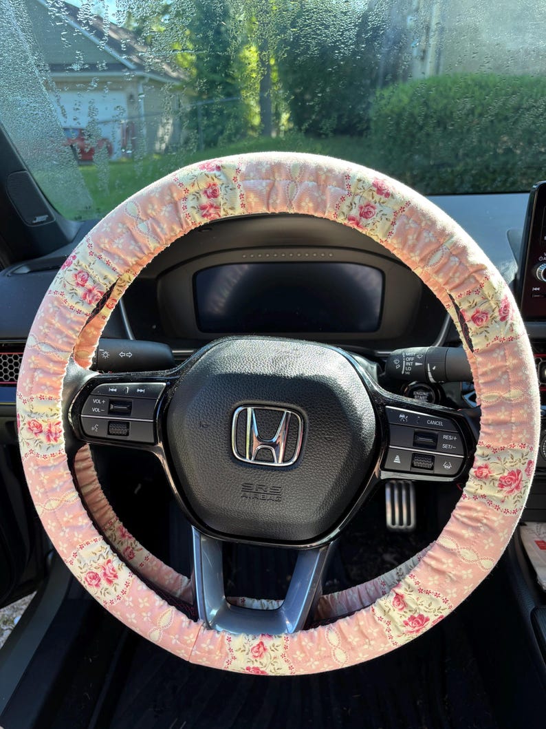Op de afbeelding: Een roze bloemen stuurwielhoes op een zwart stuurwiel. De hoes heeft een herhalend patroon van roze rozen en groene bladeren op een lichtroze achtergrond. Het dashboard en de bedieningselementen van de auto zijn zichtbaar.