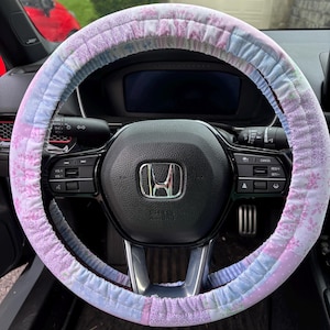 May include: A fabric steering wheel cover featuring a patchwork design with various shades of light purple, pink, and blue floral and polka dot patterns. The cover is gathered with elastic and fitted around a black Honda steering wheel with controls.