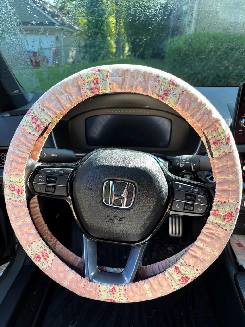 Op de afbeelding: Een roze bloemen stuurwielhoes op een zwart stuurwiel. De hoes heeft een herhalend patroon van roze rozen en groene bladeren op een lichtroze achtergrond. Het dashboard en de voorruit van de auto zijn zichtbaar op de achtergrond.