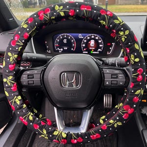 May include: A black steering wheel cover with a cherry pattern. The cover features red cherries, green leaves, and white polka dots on a black background. The steering wheel is black with various controls and a silver emblem.