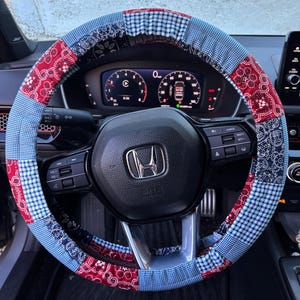 May include: A patchwork steering wheel cover in red, white, and blue. The cover is made of fabric squares in paisley, gingham, and solid patterns. The cover is on a black steering wheel with a Honda logo.