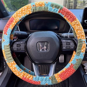 May include: A fabric steering wheel cover featuring a patchwork design of floral patterns in orange, light blue, and olive green. The cover is fitted over a black Honda steering wheel with integrated control buttons and a central silver logo emblem.