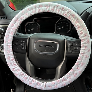 May include: A white steering wheel cover with a repeating pattern of pink bows. The cover is installed on a black steering wheel in a vehicle. The dashboard and other interior components are visible in the background.