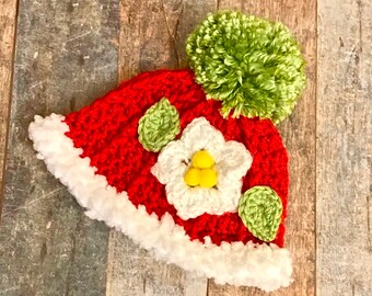 May include: Red crocheted hat with a white flower and green leaves. The hat has a white fur trim and a green pom-pom on top.