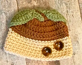Puede incluir: Un gorro de punto marrón, verde y crema con dos botones marrones. El gorro tiene un diseño único con un borde curvo y una parte superior puntiaguda.