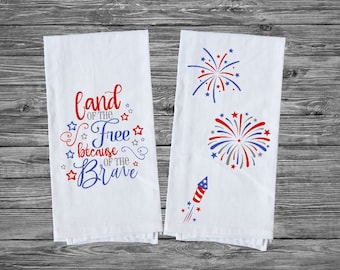 May include: Two white kitchen towels with red, white, and blue designs. One towel features the text "Land of the Free because of the Brave" with stars. The other towel has two red, white, and blue fireworks and a red, white, and blue firework rocket.