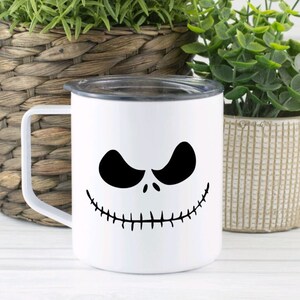 Może przedstawiać: Biały kubek ceramiczny z czarno-białym wzorem twarzy Jacka Skellingtona. Kubek ma czarną pokrywkę i uchwyt.