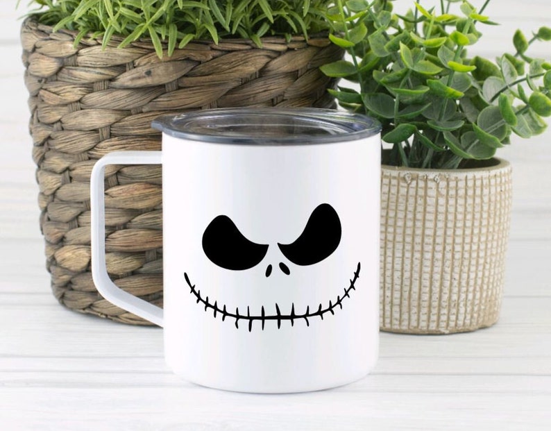 Może przedstawiać: Biały kubek ceramiczny z czarno-białym wzorem twarzy Jacka Skellingtona. Kubek ma czarną pokrywkę i uchwyt.