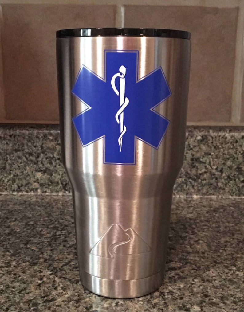 May include: Stainless steel travel mug with a blue star of life and a white caduceus medical symbol. The mug has a silver mountain logo on the bottom.