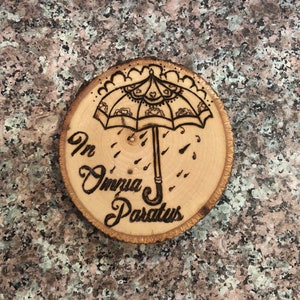 May include: A wooden round disc with a burnt design of an umbrella with raindrops and the text "In Omnia Paratus".