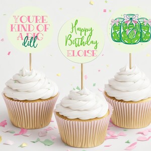 Puede incluir: Tres cupcakes con envoltorios rosas y dorados. Cada cupcake tiene glaseado blanco y un adorno de papel redondo y colorido. Los adornos dicen "You're kind of a big dill", "Happy Birthday Eloise", y presentan una imagen de dibujos animados de tres frascos de pepinillos con lazos rosas.