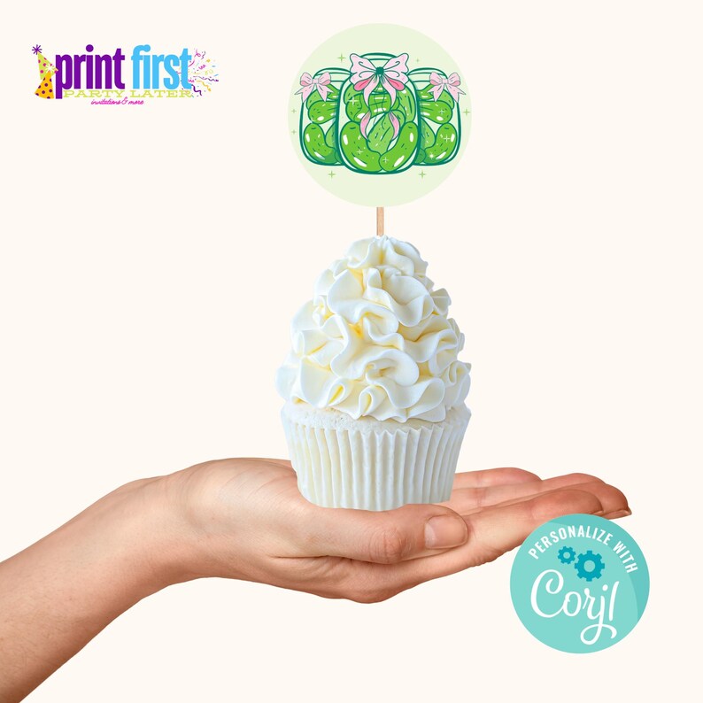 Puede incluir: Un cupcake blanco con un adorno de cupcake de tem&aacute;tica de pepinillos verde y blanco. El adorno tiene tres frascos de pepinillos con lazos rosas en la parte superior. El cupcake est&aacute; en una mano.