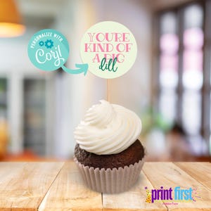 Puede incluir: Un cupcake con glaseado blanco y un topper redondo para cupcake verde claro y rosa que dice "You're kind of a big dill".
