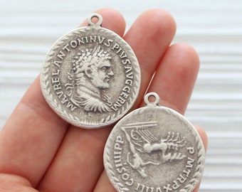May include: Two silver-toned metal pendants with Roman-inspired designs. One pendant features a profile of a man with a crown of thorns, with the text "MAVREL ANTONINV SPIVS AVG GERM". The other pendant features a winged figure with the text "PMTRP VIII COS IIII PP".
