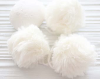 May include: Four white fluffy pom poms, three are round and one is slightly oval shaped. The pom poms are arranged on a white wooden surface.