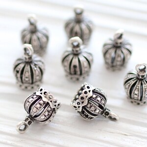 May include: A collection of eight silver-toned metal crown charms. Each charm features a detailed, textured surface with vertical ridges and a small loop at the top for attachment. The charms are arranged in a group, with some standing upright.