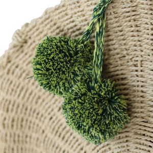 Puede incluir: Un bolso de paja tejido con pompones verdes y amarillos unidos a las asas.