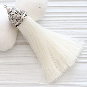 May include: A white tassel with an ornate silver metal top. The tassel is made of soft, fluffy fibres and is perfect for adding a touch of elegance to any project.