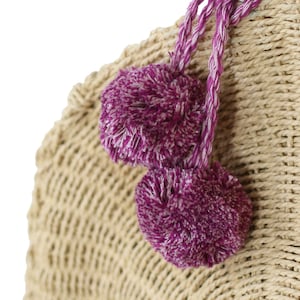 May include: A straw bag with two purple pom-poms attached to the handle. The bag is made of woven straw and has a natural color.