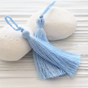 May include: Two light blue tassels with long, fluffy strands. The tassels are on a white wooden surface.