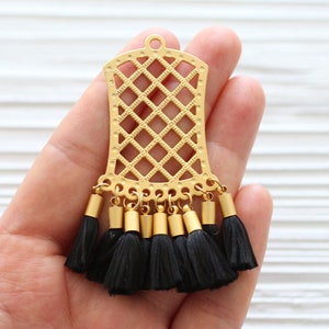 May include: A gold-colored metal pendant with a lattice design and small black tassels. The pendant has a rectangular shape with a rounded top and a small loop for hanging. The tassels are attached to the bottom of the pendant.