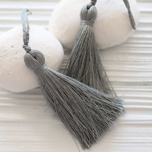 May include: Two gray tassels with looped tops and long, flowing strands. The tassels are displayed on a white, textured surface, with two white, smooth stones in the background. The tassels are a neutral color.