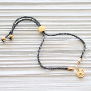 May include: A gray rope bracelet with gold-colored accents. The bracelet features a sliding knot for adjustable sizing and a small, round charm with a compass rose design. The bracelet is displayed on a white wooden surface.