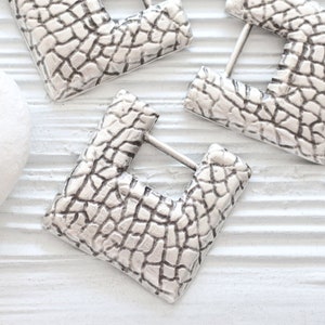 May include: Three silver square earrings with a textured surface. The earrings have a square shape with a cut-out in the center.