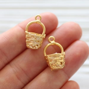 May include: Two gold-colored metal basket charms with a woven design. The charms have a loop at the top for attaching to jewelry or other crafts.
