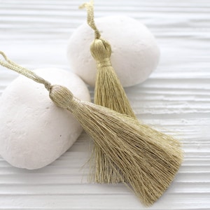 May include: Two gold-colored tassels with a silky texture are displayed against a white, textured background. The tassels are made of fine threads, with a looped top and a flowing, fringed bottom. The tassels are next to two white, smooth stones.