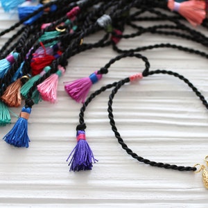 May include: A black braided cord necklace with a gold charm and a blue stone pendant. The necklace has colorful tassels attached to the cord.