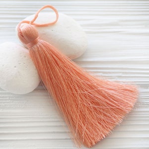May include: A peach-colored tassel with a long, fluffy fringe. The tassel is lying on a white surface.