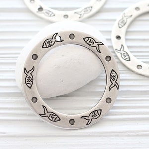 May include: Silver-toned metal circle charm with a decorative design of fish around the edge. The charm has a hole at the top for attaching to a necklace or bracelet.