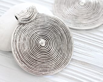 May include: Close-up of silver-toned metal pendants with a spiral design. The pendants have a circular shape with concentric lines radiating from the center. One pendant has a loop for hanging. The background is a white surface.